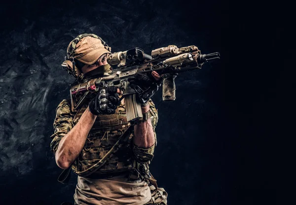 The elite unit, special forces soldier in camouflage uniform holding an assault rifle with a laser sight and aims at the target. Studio photo against a dark wall — Stock Photo, Image