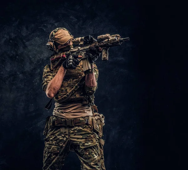 A unidade de elite, soldado das forças especiais em uniforme de camuflagem segurando um rifle de assalto com uma mira de laser e aponta para o alvo. Foto de estúdio contra uma parede escura — Fotografia de Stock