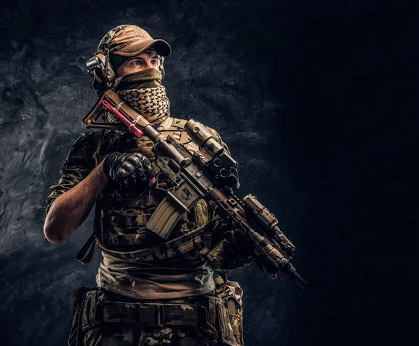 Повністю обладнаний солдат у камуфляжній формі, що тримає штурмову гвинтівку. Студійне фото на тлі темної стіни — стокове фото