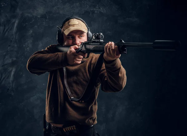 Hombre cazador en gorra y auriculares sosteniendo un rifle y apuntando a su objetivo o presa. Foto del estudio contra un fondo de pared oscuro —  Fotos de Stock