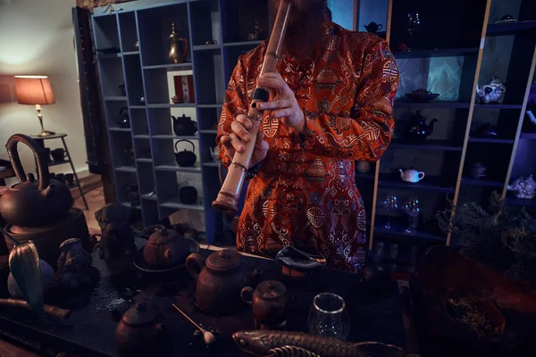 Tradition, health, harmony. Chinese tea ceremony. Tea master in kimono performs in the dark room with a wooden interior, playing on a bamboo flute — Stock Photo, Image