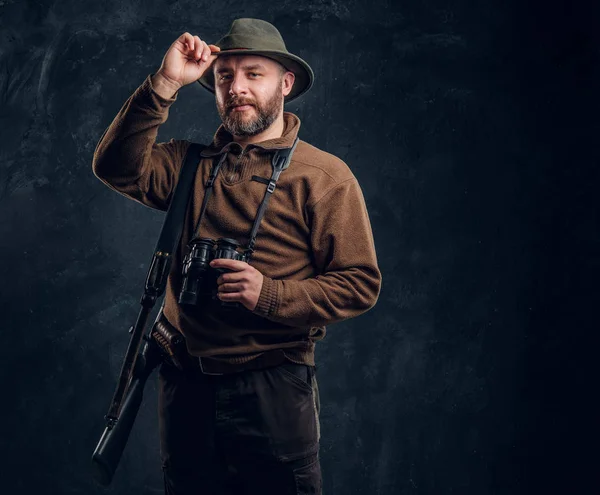 Caçador barbudo com rifle e binóculos corrige seu chapéu e olhando para uma câmera. Foto do estúdio contra um fundo de parede escura — Fotografia de Stock