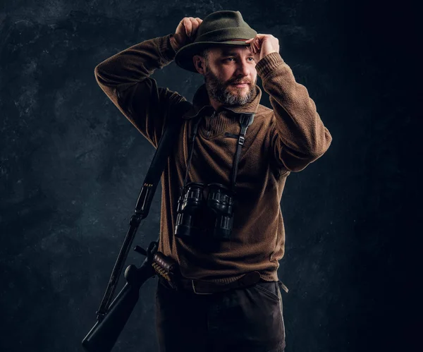 Caçador barbudo com rifle e binóculos corrige seu chapéu e olhando para o lado. Foto do estúdio contra um fundo de parede escura — Fotografia de Stock
