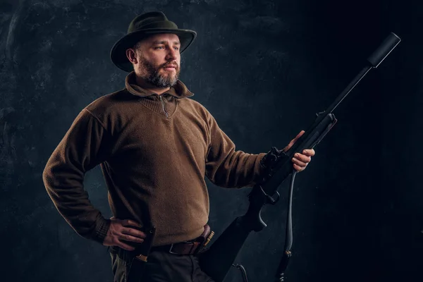 Cazador seguro posando con un rifle y mirando hacia los lados. Estudio foto contra fondo oscuro de la pared —  Fotos de Stock