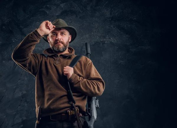 Portrait of a bearded hunter with rifle holding hand on hat and looking sideways. Studio photo against a dark wall background — Stock Photo, Image