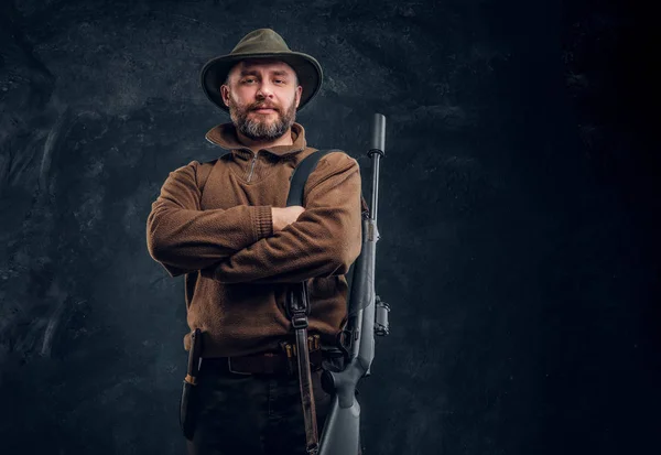 Portrait of a bearded hunter with rifle posing with his arms crossed. Studio photo against a dark wall background — Stock Photo, Image