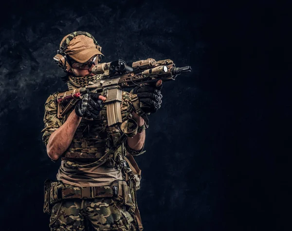 A unidade de elite, soldado das forças especiais em uniforme de camuflagem segurando um rifle de assalto com uma mira de laser e aponta para o alvo. Foto de estúdio contra uma parede escura — Fotografia de Stock