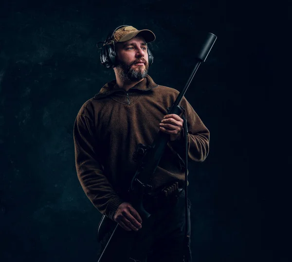 Retrato de um caçador em boné e fones de ouvido segurando um rifle e olhando para o lado. Foto do estúdio contra um fundo de parede escura — Fotografia de Stock