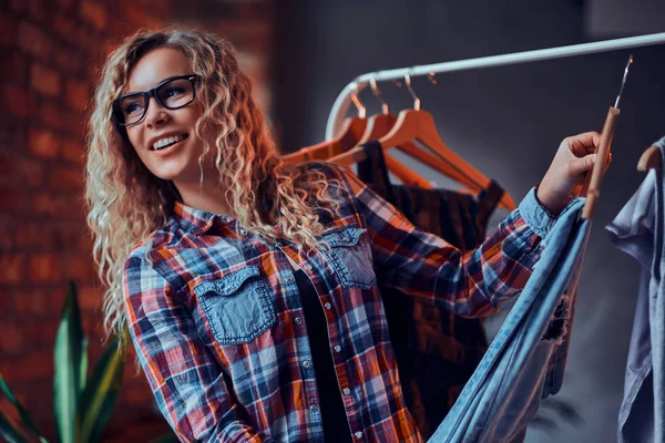 Mulher alegre sorridente em óculos e camisa quadriculada escolhe roupas em butique para suas férias — Fotografia de Stock