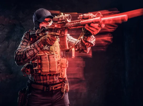 Special forces soldier wearing a checkered shirt and protective equipment holding an assault rifle and aim at the enemy. Red light effect in motion. — Stock Photo, Image