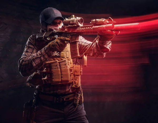 Special forces soldier wearing a checkered shirt and protective equipment holding an assault rifle and aim at the enemy. Red light effect in motion. — Stock Photo, Image