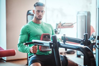 Yeşil tişörtlü genç dalgın vücut geliştirici eğitim aparatları üzerinde egzersiz yapıyor.