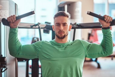 Yeşil tişörtlü genç dalgın vücut geliştirici eğitim aparatları üzerinde egzersiz yapıyor.
