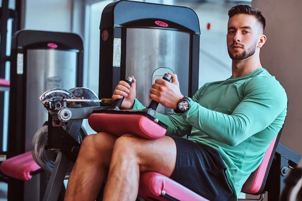 Сильный задумчивый мужчина сидит на тренировочном аппарате в спортзале и делает упражнения на ноги — стоковое фото