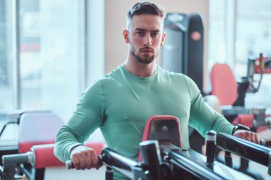 Yeşil tişörtlü genç dalgın vücut geliştirici eğitim aparatları üzerinde egzersiz yapıyor.