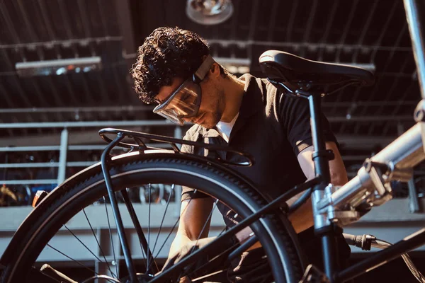 Pensive diligent master in protective glasses is fixing bicycle at workshop