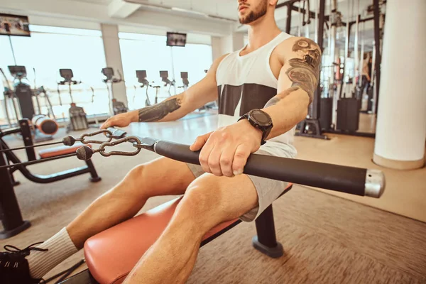 Молодой мускулистый мужчина с татуировками делает упражнения на гребной машине — стоковое фото
