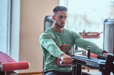 Yeşil tişörtlü genç dalgın vücut geliştirici eğitim aparatları üzerinde egzersiz yapıyor.