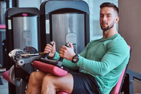 Fuerte hombre pensativo está sentado en el aparato de entrenamiento en el gimnasio y hacer ejercicios de piernas —  Fotos de Stock