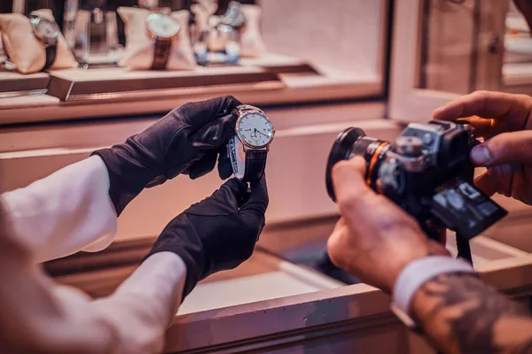 Fotógrafo tatuado está tirando foto de relógio caro na loja elegante, enquanto assistente de loja está segurando-os — Fotografia de Stock