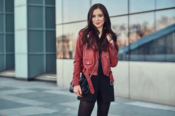 Atractiva mujer sonriente en chaqueta roja está posando cerca del edificio de vidrio —  Fotos de Stock