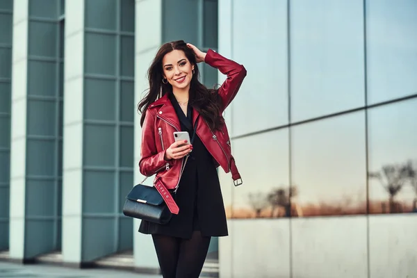 Hermosa mujer sonriente con chaqueta roja está de pie junto al edificio con teléfono inteligente en sus manos —  Fotos de Stock