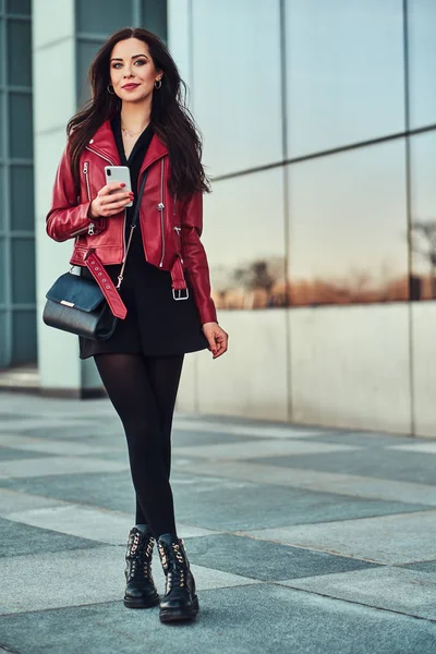 Hermosa mujer sonriente con chaqueta roja está de pie junto al edificio con teléfono inteligente en sus manos —  Fotos de Stock