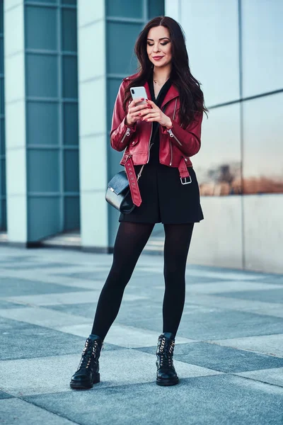 Hermosa mujer sonriente con chaqueta roja está de pie junto al edificio y chating por teléfono móvil . —  Fotos de Stock