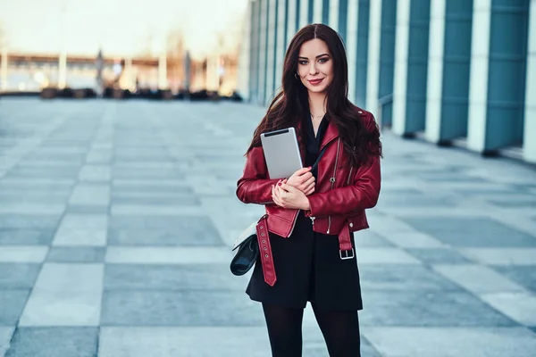 Ganska leende kvinna i röd jacka poserar nära glasbyggnad med Tablet — Stockfoto