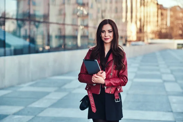 Mujer bastante sonriente en chaqueta roja está posando cerca de la construcción de vidrio con tableta —  Fotos de Stock