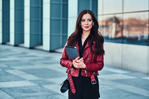 Красивая улыбающаяся женщина в красной куртке позирует возле стеклянного здания с планшетом — стоковое фото