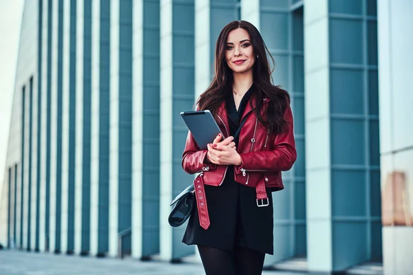 Красивая улыбающаяся женщина в красной куртке позирует возле стеклянного здания с планшетом — стоковое фото