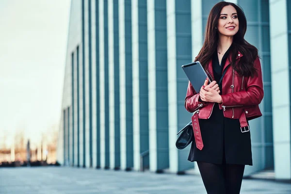 빨간 재킷을 입은 예쁜 미소 짓는 여자가 태블릿으로 유리 건물 근처에서 포즈를 취하고 있습니다. — 스톡 사진
