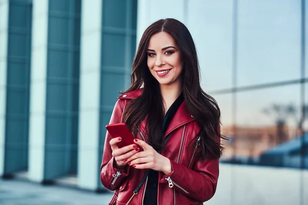 Felice donna sorridente in giacca rossa è in piedi accanto alla costruzione di vetro e chattare con qualcuno dal telefono cellulare — Foto Stock