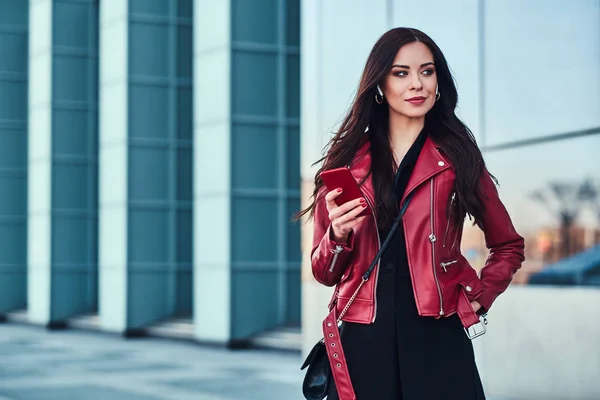 Mujer atractiva joven en chaqueta roja está disfrutando de caminar fuera mientras espera a alguien con el teléfono móvil en las manos —  Fotos de Stock