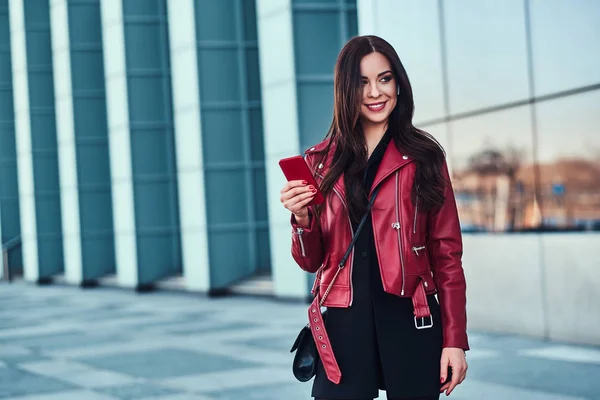 Mujer sonriente feliz en chaqueta roja está de pie junto a la construcción de vidrio y chating con alguien por teléfono móvil —  Fotos de Stock