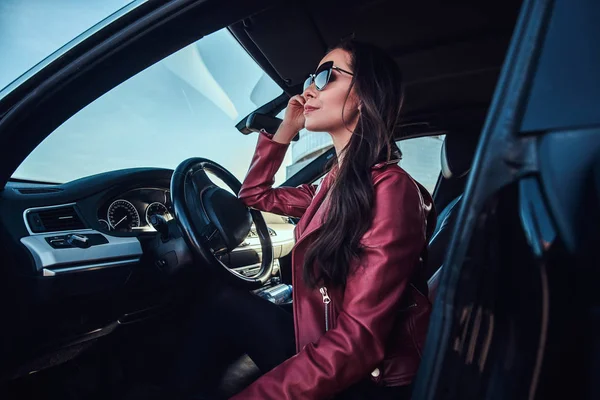 Atractiva mujer pensativa en chaqueta roja y gafas de sol está posando en su coche — Foto de Stock