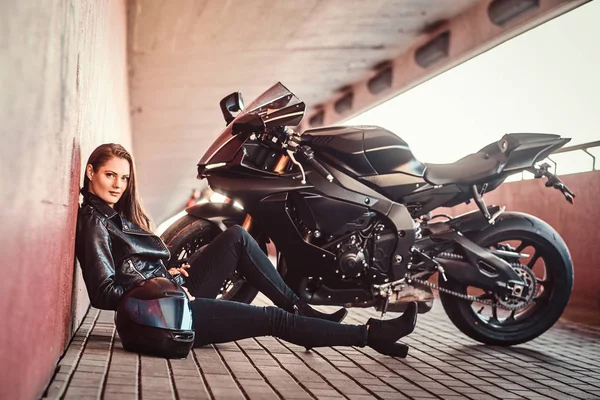 Beautiful serious woman is sitting near her bike in tunnel and leaning on wall — Stock Photo, Image