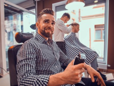 Kareli gömlekli genç adam meşgul berber dükkanında oturuyor ve elinde saç kesimi makası var.