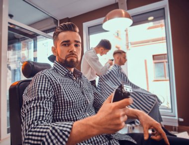 Kareli gömlekli genç adam meşgul berber dükkanında oturuyor ve elinde saç kesimi makası var.