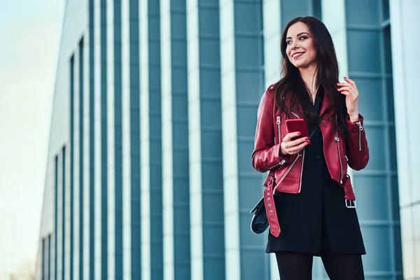 Vacker leende kvinna i röd jacka står bredvid byggnad med smartphone i hennes händer — Stockfoto