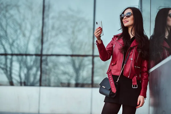 Feliz mujer sonriente en chaqueta roja y gafas de sol está disfrutando de caminar fuera mientras toma selfie en su teléfono móvil — Foto de Stock