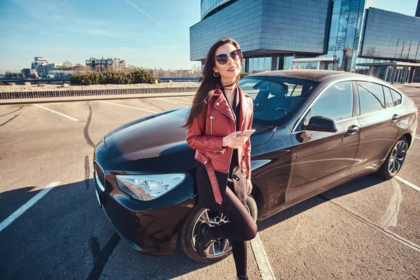 Gelukkig lachende vrouw in rode jas en zonnebril leunend op haar auto tijdens het chatten op mobiele telefoon — Stockfoto