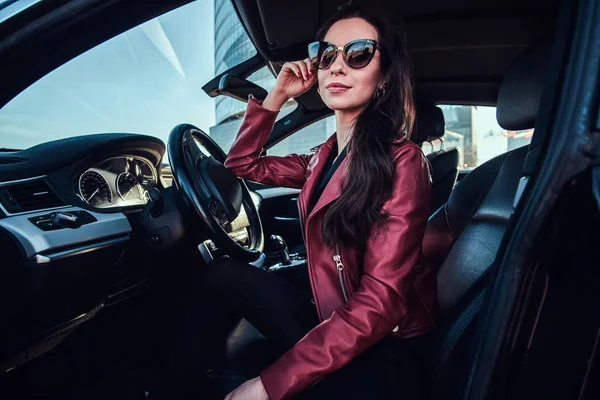 Atractiva mujer pensativa en chaqueta roja y gafas de sol está posando en su coche —  Fotos de Stock