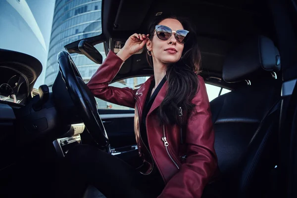 Atractiva mujer pensativa en chaqueta roja y gafas de sol está posando en su coche —  Fotos de Stock