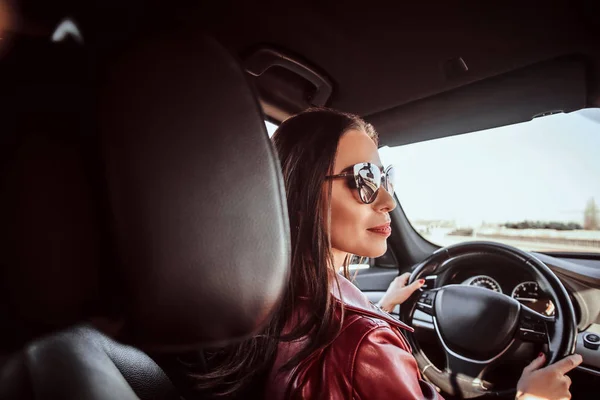 Jolie jeune femme en veste rouge et lunettes de soleil dans la conduite de sa voiture — Photo