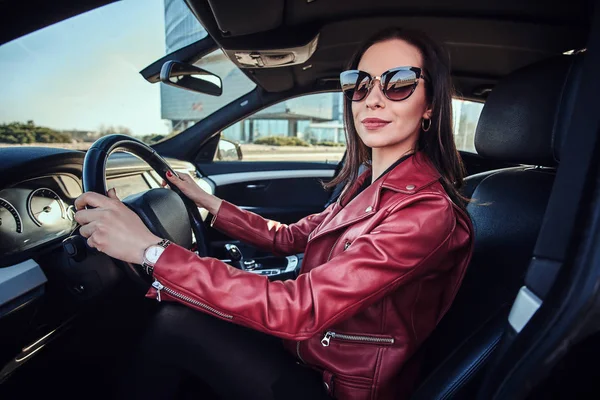 Attraktiv ung kvinna i röd jacka och solglasögon i kör sin bil — Stockfoto