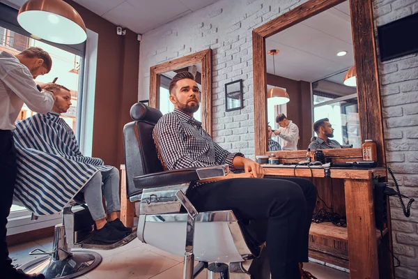 Attractive pensive man is waiting for his turn to get a haircut at busy barbershop — Stock Photo, Image