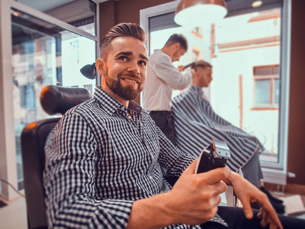 Kareli gömlekli genç adam meşgul berber dükkanında oturuyor ve elinde saç kesimi makası var.