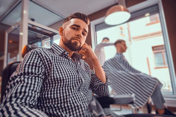 Joven estiloso hombre con camisa a cuadros está sentado en la peluquería ocupada y la celebración de trimmer para corte de pelo — Foto de Stock